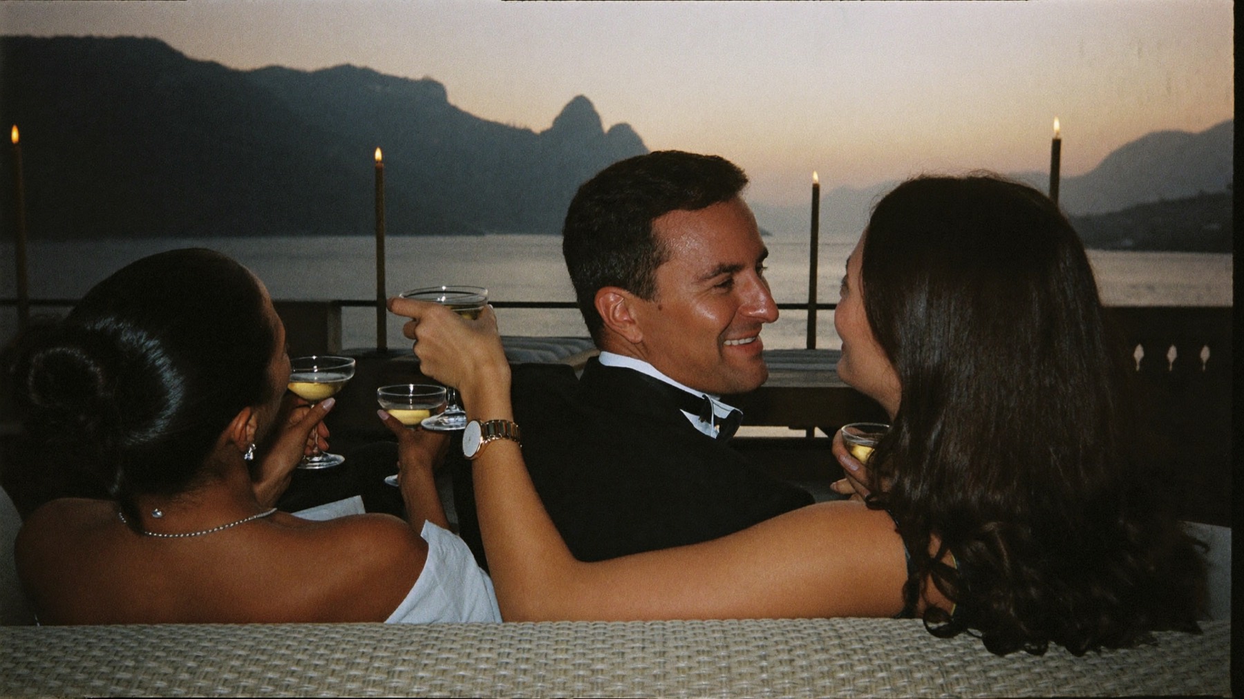 Friends in black tie raising champagne coupes on a seaside terrace at dusk
