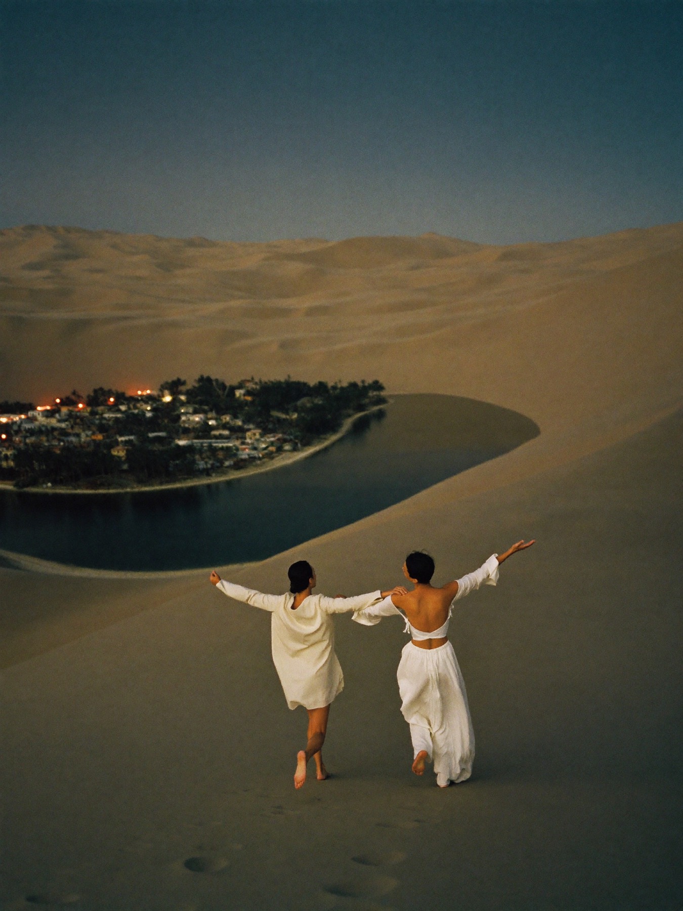 Two friends seen from behind running down a sand dune at dusk, oasis lights glowing below