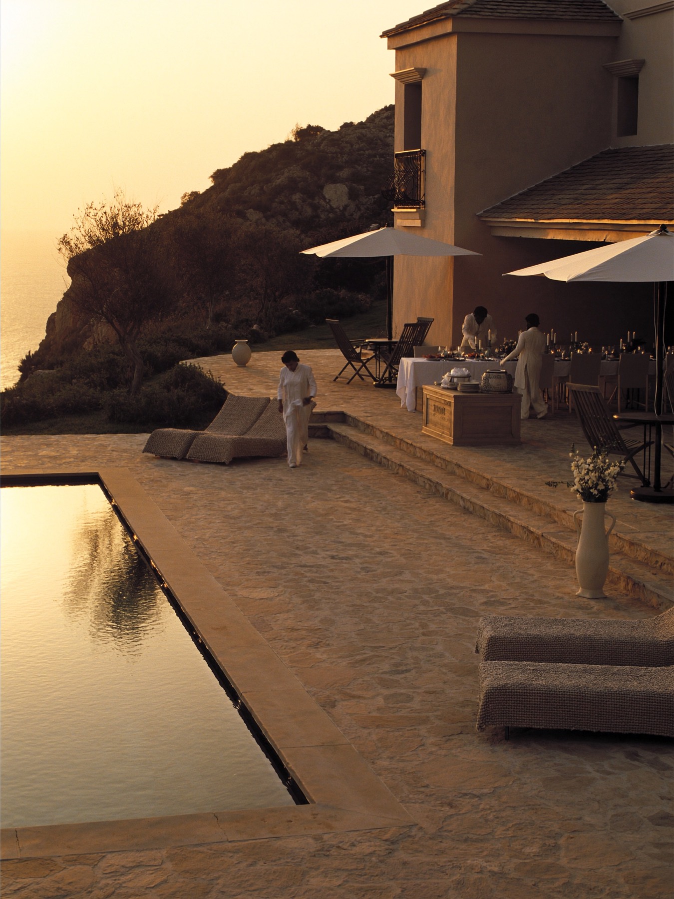 A cliffside stone estate at golden hour, staff setting a candlelit table beside the infinity pool