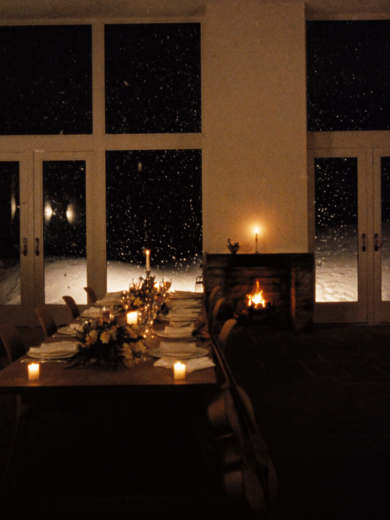 A candlelit alpine dinner table before tall windows, snow falling outside