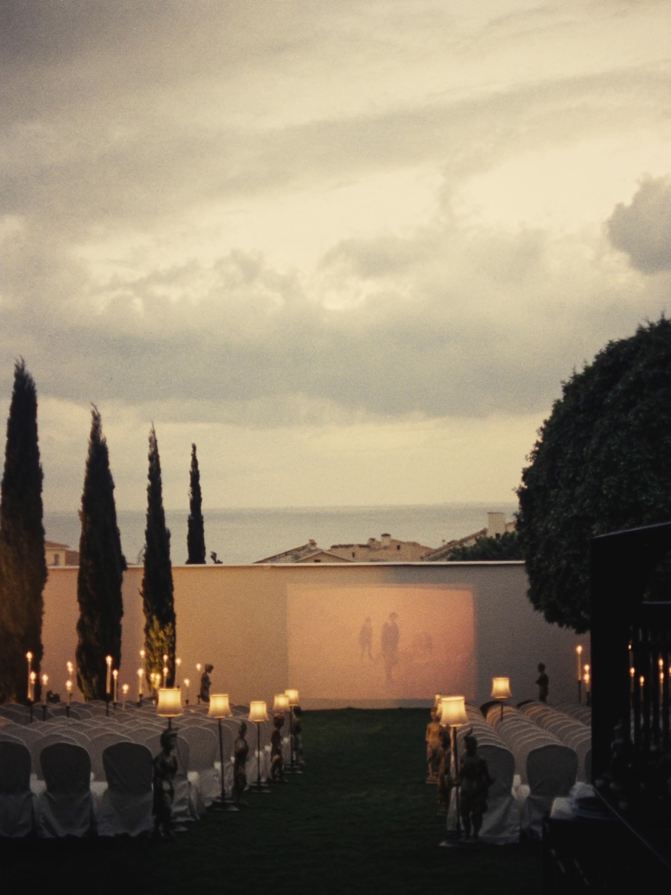 A film projected in a villa garden between cypress trees at dusk