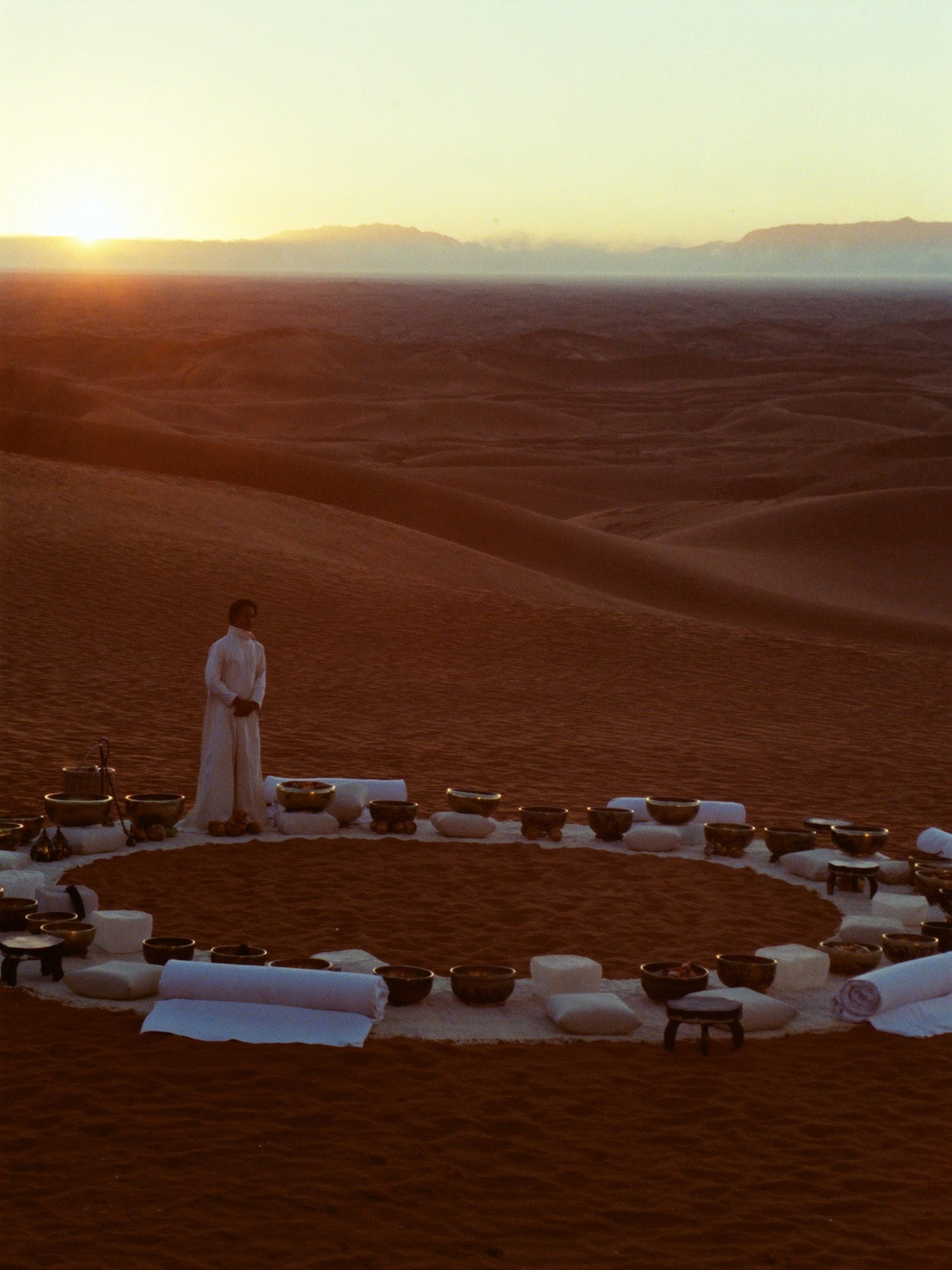 A circle of cushions and singing bowls on a red dune at sunrise